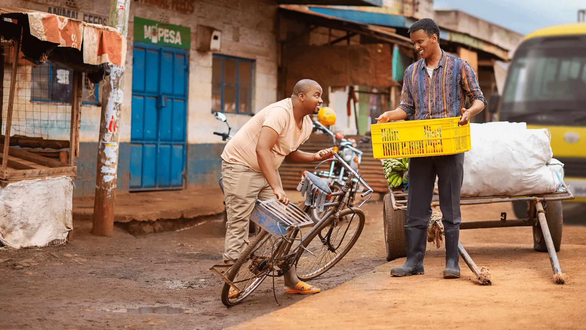 A street in Kenya with a man loading a bicycle beside a yellow crate, an M-PESA shop with a blue door behind him.